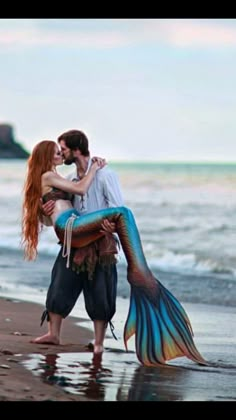 a man and woman hug on the beach while holding a mermaid tail in front of them