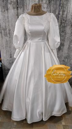 a white wedding dress with long sleeves and a bow at the neck, on display in front of a wooden wall