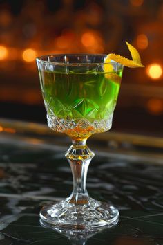 a glass filled with green liquid and topped with an orange peel garnish sitting on top of a table