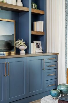 a living room with blue cabinets and white curtains on the windowsill, bookshelves and vases