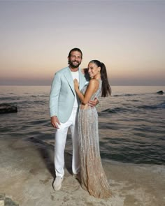 a man and woman standing on top of a beach next to the ocean at sunset