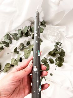 a person holding a black candle with white stars and snowflakes on it, surrounded by greenery