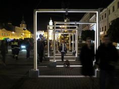 people are walking down the sidewalk in front of an open glass structure at night time