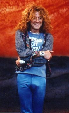 a man with long red hair standing in front of a large orange wall wearing blue pants