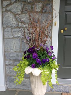 a white vase with purple and green flowers in front of a stone wall, next to a door