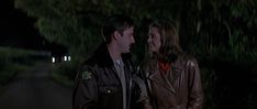 a man and woman standing next to each other on a road at night with trees in the background