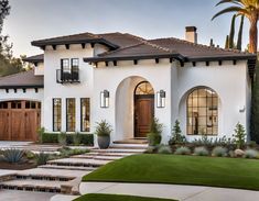 a white house with brown doors and some plants in the front yard, surrounded by palm trees