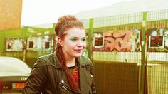 a woman with braids and green eyes stands in front of a fenced area