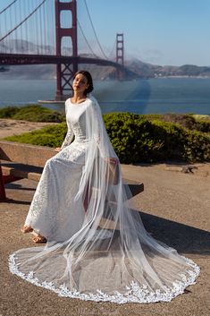 a woman wearing a white wedding gown and veil sitting on a bench in front of the golden gate bridge