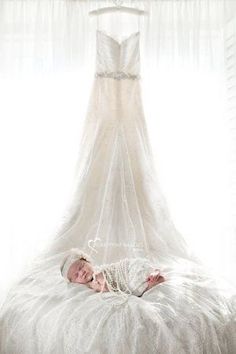 a baby laying on top of a white bed in front of a dress hanging from the ceiling