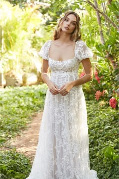 a woman in a white dress standing on a path with trees and bushes behind her