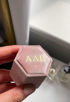 a hand holding a pink ring box with the word aa on it in gold lettering