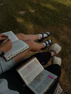 two people are sitting on the grass with open books in their lap and one is holding a dog's paw