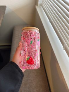 a person holding a glass jar with pink glitter and strawberries on it, in front of a window