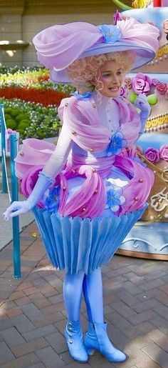 a woman dressed in pink and blue is standing next to a giant cupcake with flowers on it