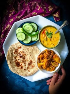 a person holding a plate with food on it and cucumbers, soup, and bread