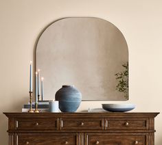 a dresser with a vase and candles on it in front of a large round mirror