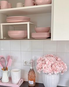 pink flowers are in a white vase on the kitchen counter next to plates and utensils