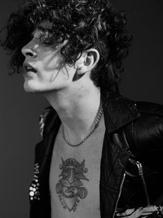 a man with curly hair wearing a leather jacket and tattoos on his chest is looking off to the side