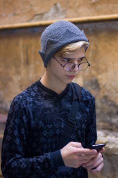 a young man wearing glasses and a hat looks at his cell phone while standing in front of a stone wall