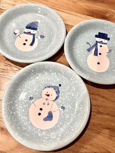 three plates with snowmen painted on them sitting on a wooden table next to each other