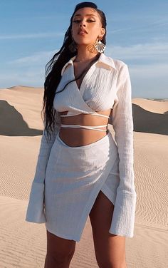 a woman standing in the sand wearing a white dress and high heeled sandals with her hands on her hips