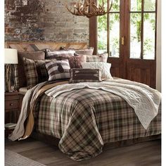 a bed with plaid comforter and pillows in front of a brick wall next to a window