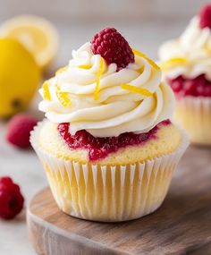 two cupcakes with white frosting and raspberries on a wooden board