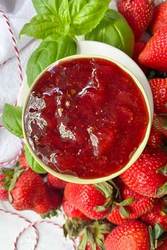 a bowl of strawberry jam surrounded by fresh strawberries on a white cloth with basil leaves