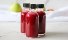 three bottles filled with red liquid sitting on top of a white counter next to an apple