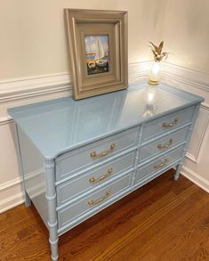 a blue dresser with gold hardware and a painting on it's top, next to a wooden floor