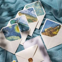 four envelopes with paintings on them sitting on a blue blanket next to a notepad