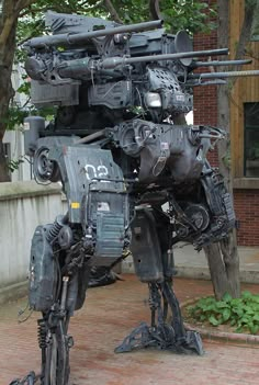 a large metal robot standing on top of a brick sidewalk next to a tree and building