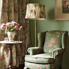 a green chair with pink flowers on it next to a lamp and flowered curtains