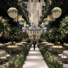 a man walking down a long aisle covered in lush greenery and hanging lanterns above tables