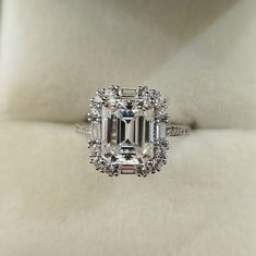 an emerald - cut diamond ring is displayed on a white cloth covered surface, surrounded by diamonds