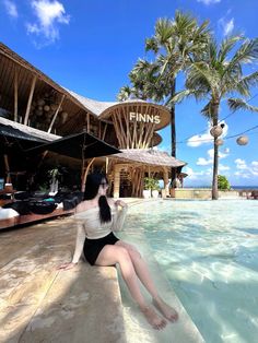 a woman sitting on the edge of a swimming pool in front of a building with palm trees