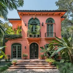 a pink house with green shutters and plants on the front steps, surrounded by palm trees