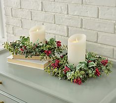 two white candles are sitting on a table with holly and berries in front of a brick wall