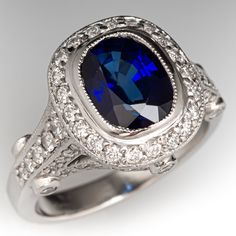 This glimmering ring is centered with one (1) bezel set cushion cut sapphire that is bordered by twenty-one (21) bead set round brilliant cut diamonds. The shoulder and side faces of the ring are set with sixty-eight (68) bead set round brilliant cut diamonds, and six (6) bezel set round brilliant cut diamonds. The ring has milgrain edging.  It measures 12.9mm at the top, rises 7.1mm above the finger, and tapers to 2.5mm wide by 1.5mm thick at the base of the shank. There are a few very light sw Cushion Cut Sapphire Diamond Ring For Formal Occasions, Formal Sapphire Cushion Cut Diamond Ring, Luxury Cushion Cut Sapphire Diamond Ring, Luxury Sapphire Ring With Cushion Cut Center Stone, Luxury Cushion Cut Sapphire Ring With Center Stone, Formal Cushion Cut Sapphire Ring With Diamond, Luxury Gia Certified Cushion Cut Sapphire Ring, Platinum Sapphire Ring With Diamond Accents, Cushion Cut, Luxury Cushion Cut Sapphire Ring With Halo Setting