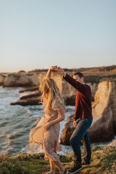 a man and woman dancing near the ocean with cliffs in the background during their engagement photo session