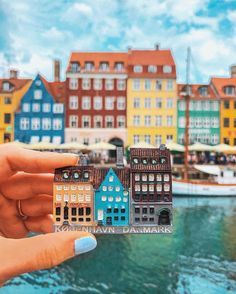 a person holding up a miniature house in front of some buildings on the water's edge