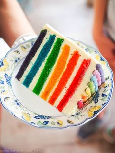 a slice of rainbow cake on a plate with someone's hand holding it in the background