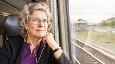 an older woman wearing headphones looking out the window at train tracks and grass in the background