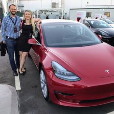 a man and woman standing next to a red tesla car in a parking lot with other cars