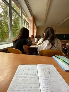 two women sitting at a table with notebooks and papers on it, in front of large windows