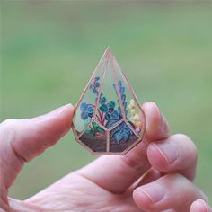 a hand holding a small piece of art with flowers on it's side and trees in the background