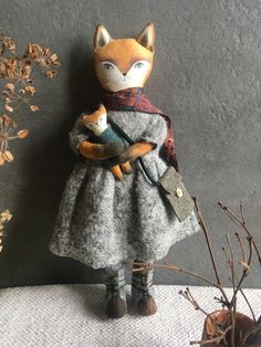 a stuffed fox is holding a cat in a gray dress and scarf on a table