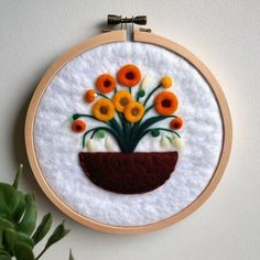 an embroidery project with orange and white flowers in a brown pot on a wall next to a green plant