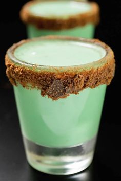 two shot glasses filled with green liquid and some brown sugar on the rim, sitting on a black surface